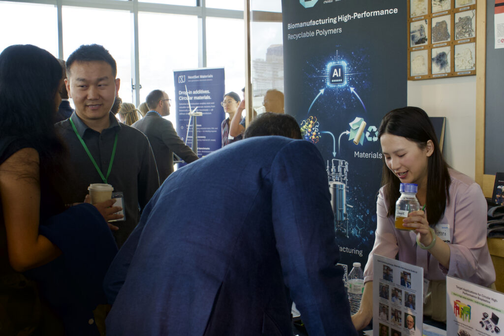 FutureBio CEO Zilong Wang smiles at an attendee on the far left while FutureBio Research Associate Weixi Hu talks to an attendee on the right, holding up a clear glass bottle of amber liquid that demonstrates FutureBio's process.