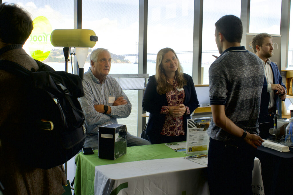 Root Applied Sciences CCO Dion Harste sits behind a table and CEO Sarah Placella stands next to him as both talk to an attendee.