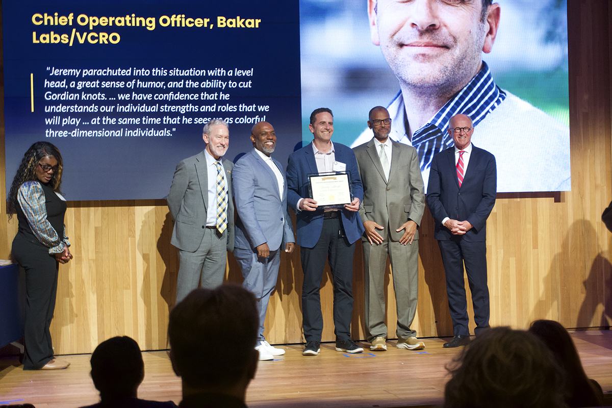 A group of 5 men in suits pose in a row, smiling, on a brightly lit stage. The man in the center, wearing a lapis blue sports coat, is holding a certificate encased in a clear glass frame. Above them, a large projector screen shows the lower half of the man's face (he is the award recipient), and text reading "Chief Operating Officer, Bakar Labs/VCRO. Jeremy parachuted into this situation with a level head, a great sense of humor, and the ability to cut Gordian knots... we have confidence that he understands our individual strengths and roles that we will play...at the same time that he sees us as colorful, three-dimensional individuals." To the left of the stage, a woman stands with her hands clasped at waist height.