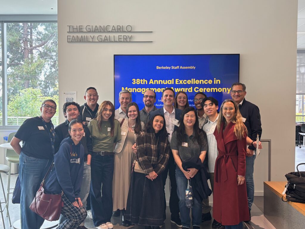 A group of 16 people poses together, smiling, in front of a large television screen that reads: "Annual Excellence in Management Award Ceremony."